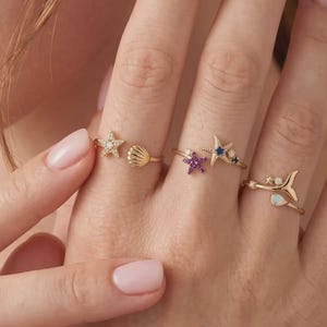 Puede incluir: Tres anillos abiertos de tono dorado con motivos marinos. Un anillo tiene una estrella de mar y una concha. El segundo anillo muestra una estrella de mar morada y una dorada con piedras azules. El tercer anillo presenta una cola de ballena.