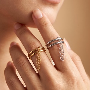 Op de afbeelding: Twee dubbele ringen, één in een goudkleurige en één in een zilverkleurige afwerking, elk voorzien van een delicaat bungelend kettinkje bevestigd aan de bovenste ring. De ringen worden om twee afzonderlijke vingers gedragen.