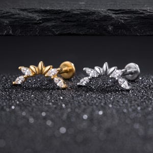 Puede incluir: Dos pendientes tipo labret con un diseño curvo en forma de abanico adornado con piedras transparentes de corte marquesa. Uno tiene un acabado en tono dorado pulido y el otro en tono plateado pulido, ambos con una base circular plana.