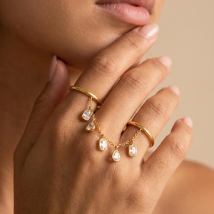 Könnte beinhalten: Ein goldfarbener Doppelring, der durch eine feine Kette verbunden ist, an der fünf klare, facettierte Steine in verschiedenen Formen, darunter rechteckige und tropfenförmige Schliffe, zwischen zwei glatten Metallbändern hängen.
