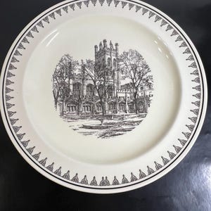 May include: A cream-colored ceramic plate featuring a detailed black ink illustration of a gothic-style building with a tall tower and trees in the center. The rim is decorated with a repeating black geometric pattern of pointed arches.