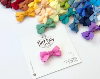 Könnte beinhalten: Eine rosa Haarschleife aus Ripsband auf einer weißen Präsentationskarte mit dem Text Tiny Paw Club Hair Accessories und your fur-baby's new favorite hair clips. Im Hintergrund ist eine Sammlung verschiedenfarbiger Haarschleifen verstreut.