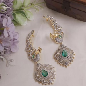 May include: A pair of gold-toned ear cuffs featuring a peacock motif encrusted with clear white crystals and a central teardrop-shaped green stone. The design includes intricate metalwork and a dangling pendant element with a matching green stone.