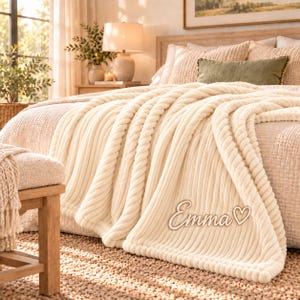 Pode incluir: Uma manta de malha grossa na cor creme com uma textura semelhante a uma corda, drapeada sobre uma cama. A manta apresenta o nome Emma seguido de um símbolo de coração numa fonte cursiva bordada ou aplicada no canto inferior direito.