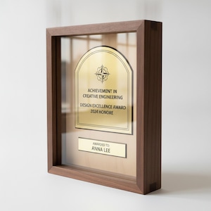 Puede incluir: Una placa de reconocimiento rectangular de madera con frente de cristal y una placa de metal dorado. La placa muestra un icono de brújula, el texto ACHIEVEMENT IN CREATIVE ENGINEERING, DESIGN EXCELLENCE AWARD 2024 HONORE y AWARDED TO: ANNA LEE.