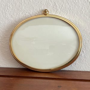 May include: An oval-shaped picture frame with a polished gold-tone metal border and a clear glass front. The frame features a small, circular hanging loop at the top center, designed for wall mounting. The interior is empty, showing a white background.