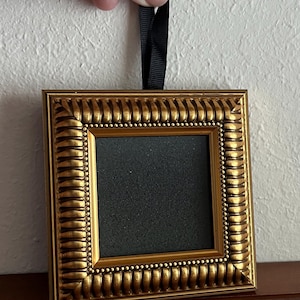 May include: A square picture frame with a metallic gold finish featuring a textured, ribbed border and a beaded inner edge. The center of the frame is filled with a solid black square insert. A black fabric ribbon is attached to the top for hanging.