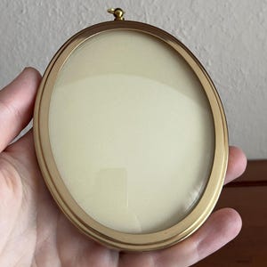 May include: An oval-shaped picture frame with a smooth, polished gold-tone metal finish. The frame features a clear glass front covering a plain, cream-colored insert and includes a small, circular gold-tone hanging loop at the top.