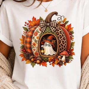 May include: A white t-shirt featuring a large graphic of a pumpkin with varied patterns including plaid, floral, and leopard print. A small ghost wearing a witch hat and glasses holds a lantern inside the pumpkin, surrounded by autumn leaves, berries, and mushrooms.