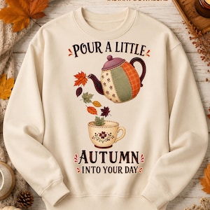 May include: A cream-colored crewneck sweatshirt featuring a graphic of a patchwork teapot pouring colorful autumn leaves into a teacup. The text reads "POUR A LITTLE AUTUMN INTO YOUR DAY" in dark brown lettering. The design includes various fall foliage.