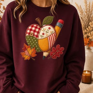May include: A dark maroon long-sleeve sweatshirt featuring a central applique design of a patchwork apple and a pencil. The apple is composed of red gingham, green polka dot, and floral fabric segments with a small wooden button. Two autumn leaves flank the apple.