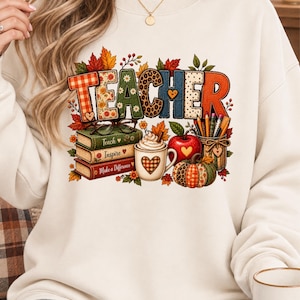 Puede incluir: Una sudadera de cuello redondo color crema con un estampado gráfico que muestra la palabra TEACHER en letras coloridas con patrones. Debajo hay tres libros apilados con las etiquetas Teach, Inspire y Make a Difference, una taza con crema batida, una manzana y una calabaza.