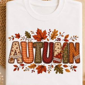 Może przedstawiać: Biała bluza z okrągłym dekoltem z napisem AUTUMN wykonanym dużymi literami. Każda litera ma unikalny wzór w stylu patchwork w odcieniach pomarańczu, czerwieni, zieleni i brązu, ozdobiony małymi guzikami, koronką i jesiennymi liśćmi.