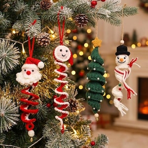 May include: Four crocheted Christmas ornaments hanging from a tree. The ornaments include a Santa Claus, a round face with a red and white spiral, a green Christmas tree with small white beads, and a snowman wearing a black hat and scarf.