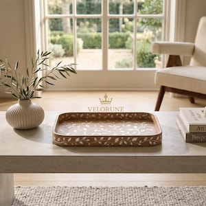May include: A rectangular wooden tray featuring an intricate white inlaid leaf pattern across its surface. The tray is positioned on a light-colored table next to a ribbed cream-colored vase with olive branches and books labeled Velorune.