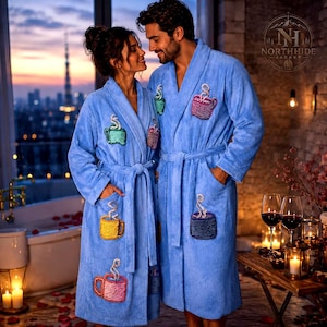 Peut inclure: Deux peignoirs assortis en tissu bleu clair doux, ornés de motifs de tasses à café brodées dans des tons jaune, rose, vert et violet. Chaque peignoir présente un col châle, des manches longues et une ceinture à nouer à la taille.