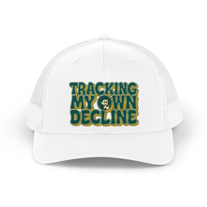 May include: A white trucker cap featuring a graphic print with the text TRACKING MY OWN DECLINE in bold, dark green letters with a yellow outline. The design includes a small, circular illustration of a cartoon character in the centre.