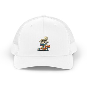 Pode incluir: Um boné trucker branco com uma ilustração gráfica de um esqueleto vestindo calças de ganga azuis e a andar num triciclo vermelho, amarelo e azul. O boné tem um painel frontal sólido e um painel traseiro em rede, com uma aba curva.