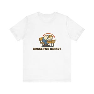 Op de afbeelding: Een wit t-shirt met een cartoonillustratie van een figuur die het hoofd op een schoolbankje laat rusten, met een rode stippellijn die beweging aangeeft. Onder de afbeelding staat de tekst BRACE FOR IMPACT in dikke bruine hoofdletters.