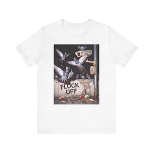 Puede incluir: Una camiseta blanca con un estampado gráfico de varias palomas volando alrededor de una cámara de seguridad montada en un poste. Debajo de las aves, el texto FLOCK OFF está estarcido en negro sobre un muro de hormigón con escombros.