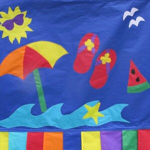 May include: A blue fabric banner with a yellow sun wearing sunglasses, a red and yellow beach umbrella, a blue wave, a yellow star, and red flip flops. The banner has a watermelon slice and a row of colorful fabric strips at the bottom.