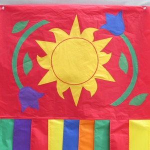 May include: A red fabric wall hanging with a yellow sun, blue and purple flowers, and green leaves. The bottom of the hanging has vertical stripes in rainbow colors. The wall hanging is attached to the wall with two white push pins.