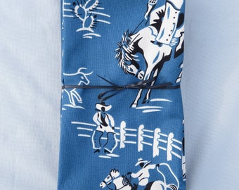 May include: A stack of blue cotton napkins with a white cowboy and horse design. The napkins feature a repeating pattern of cowboys riding horses, cacti, and fences.