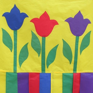 May include: Three colorful felt tulips with green leaves on a yellow background. The tulips are blue, red, and purple. The bottom of the image shows a row of colorful stripes.