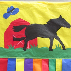 May include: A yellow fabric banner with a red barn, a black silhouette of a horse running, and a blue cowboy hat. The banner has a green grass border and a rainbow fringe.