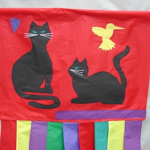May include: Red fabric banner with two black cat silhouettes and a yellow bird silhouette. The banner has a purple, yellow, red, green, and purple fringe.