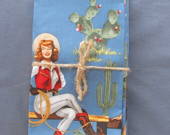 May include: A set of four blue cotton napkins with a vintage western design featuring a woman in a cowboy hat and boots, cacti, and a rope. The napkins are tied together with twine.