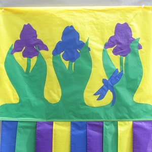 May include: A yellow fabric banner with three purple, blue, and green iris flowers. The banner has a green border and a yellow, purple, and blue striped bottom.
