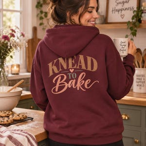 May include: A deep maroon hooded sweatshirt featuring the text KNEAD TO BAKE printed on the back in a mix of tan and muted green lettering, accented by a small heart icon. The hoodie includes a hood and long sleeves, shown from the back view.