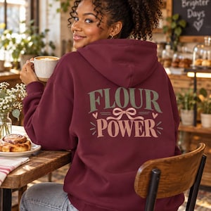 May include: A maroon hooded sweatshirt featuring the text "FLOUR POWER" printed on the back in sage green and light pink. The design includes a small bow graphic between the words and two small hearts on either side of the bow.