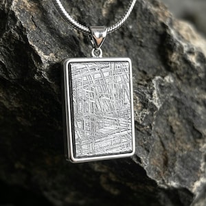May include: A rectangular pendant featuring a metallic surface with a distinct cross-hatched Widmanstätten pattern, encased in a polished silver-toned frame. The pendant hangs from a silver-toned snake chain necklace, showcasing a textured, dark gray finish.
