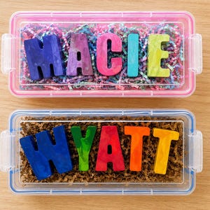 May include: Two clear rectangular plastic containers holding custom letter-shaped crayons. The top container holds the name MACIE in purple, pink, and green crayons on colorful paper shreds. The bottom container holds WYATT in blue, green, red, orange, and yellow crayons on brown paper shreds.