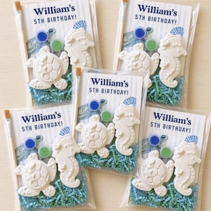May include: Five clear plastic favor bags, each containing a white ceramic turtle and seahorse figurine, a wooden paintbrush, two small paint pots in blue and green, and blue shredded paper, with a card reading William's 5TH BIRTHDAY!