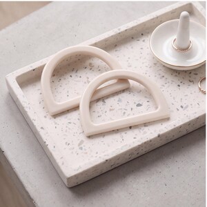 May include: A rectangular tray with a light pink and grey speckled terrazzo pattern, featuring two smooth, semi-circular, cream-colored handles resting on its surface, alongside a small white ring holder with a gold rim and a thin metal ring.