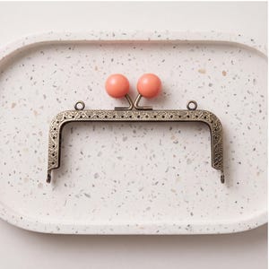 May include: A rectangular metal purse frame with an antique bronze finish and intricate embossed floral patterns along the edges. The closure features two round, smooth, coral-colored beads that act as the clasp mechanism for opening and closing.