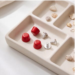 May include: Three small, glossy red conical knobs with a smooth finish, accompanied by three matching silver-toned metal screws with flat heads and threaded shafts, arranged together on a light beige, multi-compartment rectangular tray.