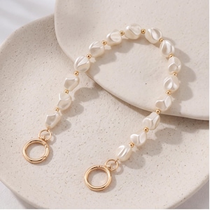 Op de afbeelding: Een decoratieve tasriem met een reeks onregelmatige, gebroken witte, parelachtige kralen afgewisseld met kleine, ronde, goudkleurige metalen tussenstukjes. De riem eindigt aan beide uiteinden met een ronde, goudkleurige metalen veerringsluiting.