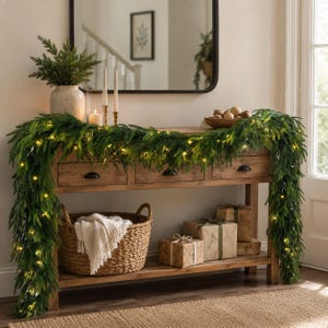 May include: A wooden console table featuring three drawers, draped with a green pine garland illuminated by warm string lights. A woven basket with a white throw, wrapped gift boxes, a ceramic vase with greenery, and candles are displayed.