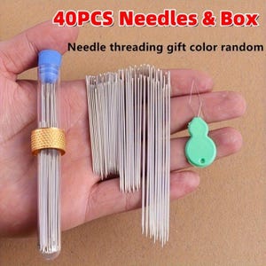 May include: A set of 40 silver sewing needles, a clear plastic storage tube with a blue cap and gold-colored metal band, and a light green gourd-shaped needle threader. The text 40PCS Needles & Box and Needle threading gift color random is visible.