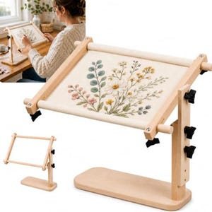May include: A light-colored wooden embroidery stand featuring an adjustable frame holding a piece of fabric with a floral design. The stand includes black knobs for height and angle adjustments, a rectangular base, and a horizontal roller system.