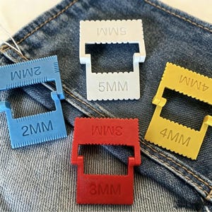 May include: Four plastic seam guides in white, blue, red, and yellow, each featuring a serrated edge and embossed text indicating sizes 2MM, 3MM, 4MM, and 5MM. These tools are designed for precise fabric alignment and consistent seam spacing.