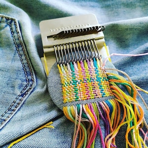 May include: A metal darning loom tool positioned on blue denim fabric, featuring a series of vertical metal hooks and a woven square patch created with multicoloured embroidery threads in shades of pink, yellow, orange, green, and blue.
