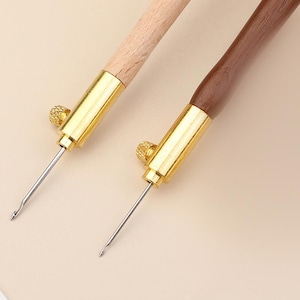 May include: Two embroidery needles with wooden handles, one light and one dark brown. Each needle features a gold-colored metal ferrule with a small, adjustable knurled screw and a thin, silver-toned metal hook at the tip for fine needlework.