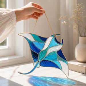 Puede incluir: Un atrapasoles de vidrieras con forma de raya manta, compuesto por un mosaico de piezas de vidrio texturizado en tonos azul oscuro, turquesa, azul claro y blanco. La pieza está delineada con una fina soldadura de metal dorado y cuelga de una cadena.