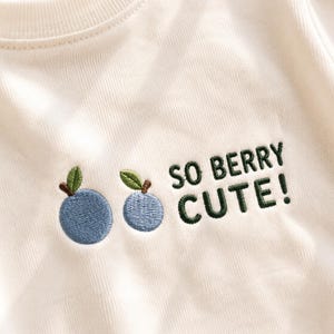 Puede incluir: Una sudadera de cuello redondo en color blanco roto con un diseño bordado de dos bayas azules con hojas y tallos verdes, acompañado por el texto bordado en verde oscuro que dice SO BERRY CUTE! en letras mayúsculas sin remates.