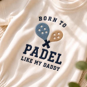 May include: An off-white short-sleeved bodysuit featuring dark blue embroidered text that reads BORN TO PADEL LIKE MY DADDY, accompanied by two crossed padel rackets in light blue and tan with circular cutouts centered on the chest.